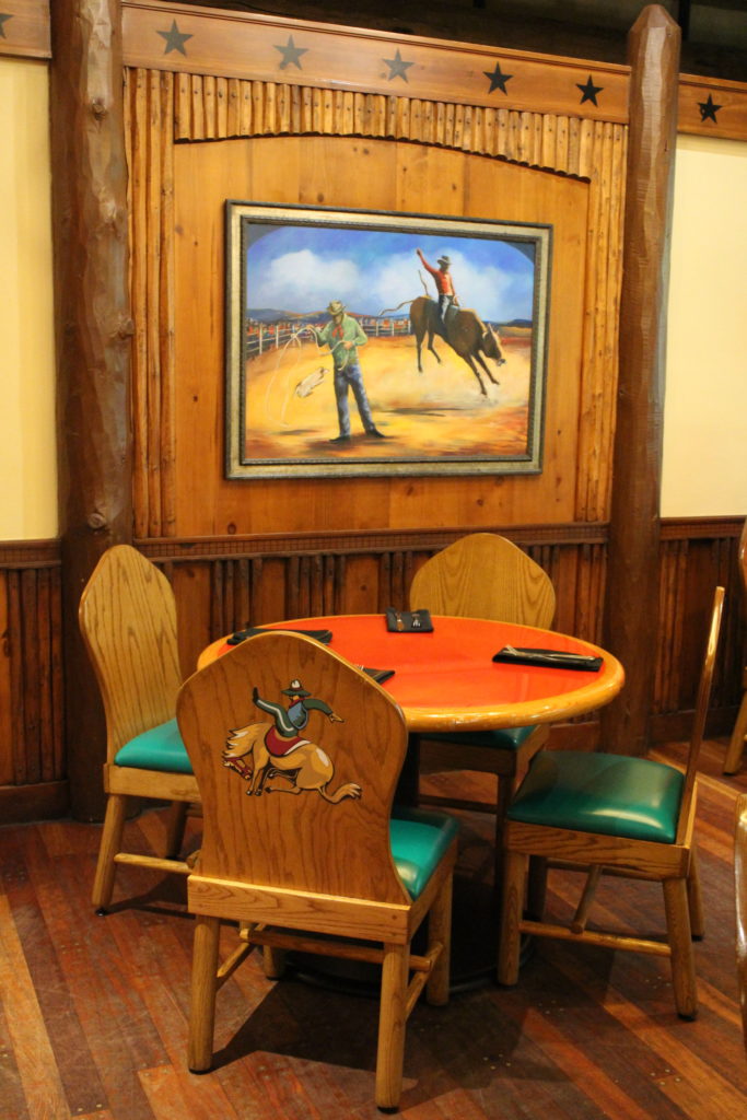 A wooden chair and tables in front of cowboy art in a western style restaurant called Whispering Canyon Cafe at Disney's Wilderness Lodge Resort