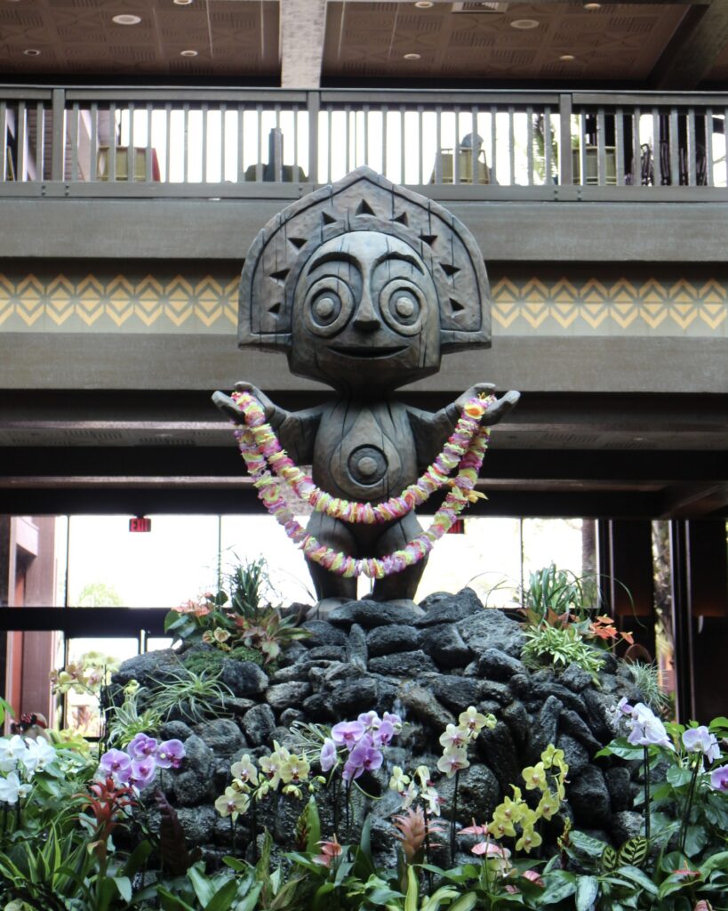 A tiki statue holds up a lei on top of a rock surrounded by flowers in Disney's Polynesian Village Resort lobby