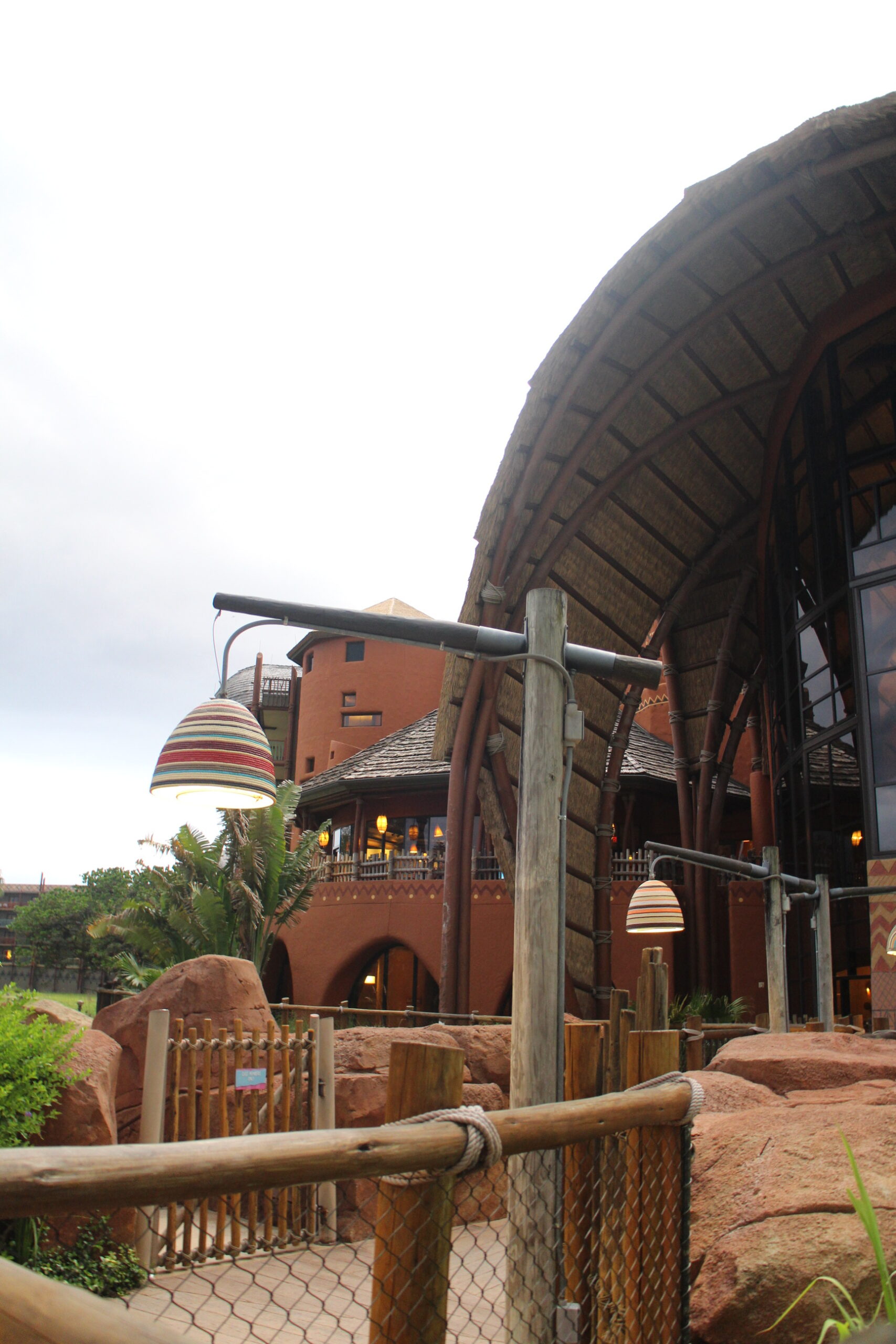 Exterior of Animal Kingdom Lodge's Kidani Village area with clay colored rock work, wood stick fences and tall windows of the lobby.