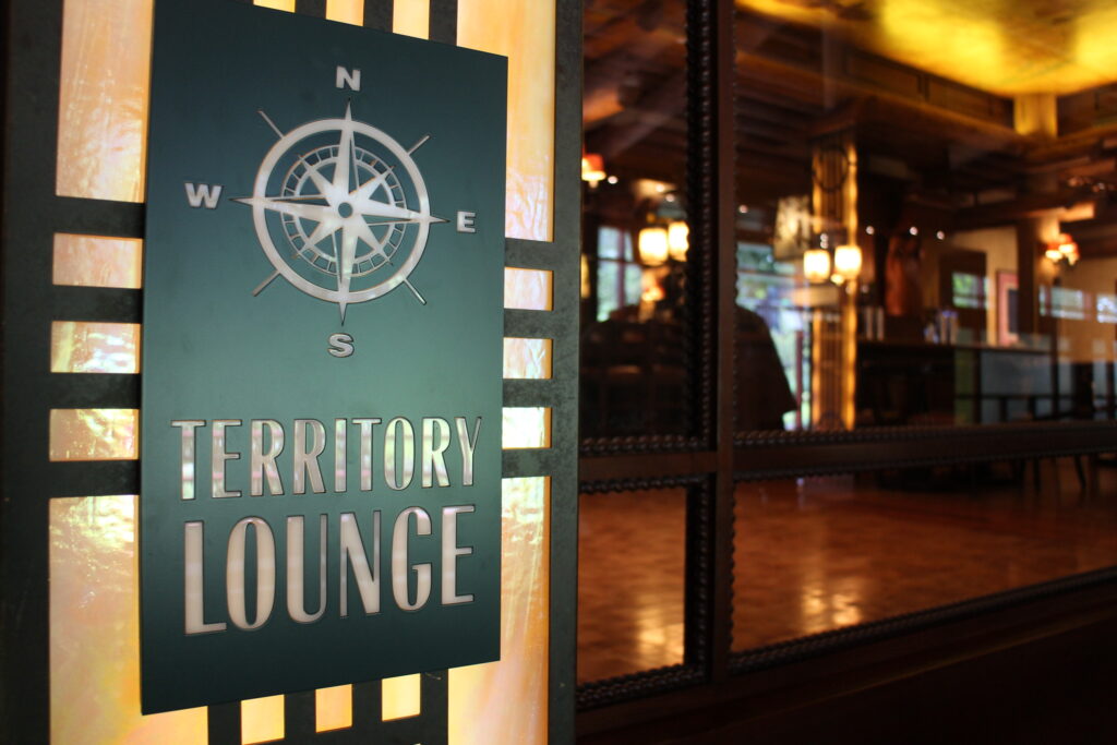 Wilderness Lodge Restaurants include Territory Lounge bar and lounge with woodsy, rustic interior behind the green, craftsman inspired Territory Lounge sign that features a compass design
