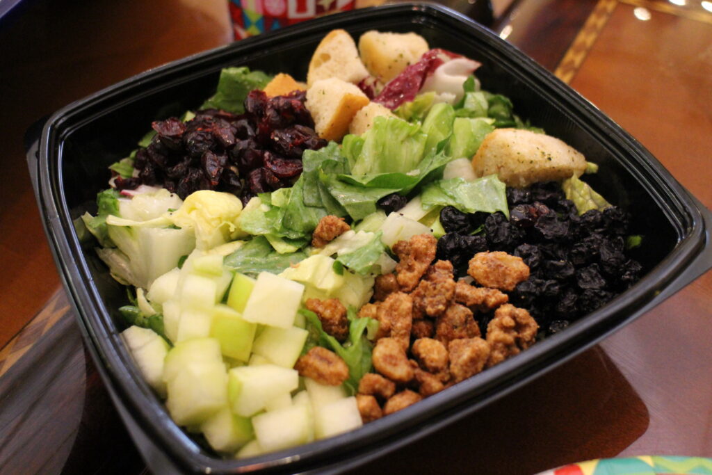 A salad with cranberries, cut up green apples, nuts and croutons in a black to go container