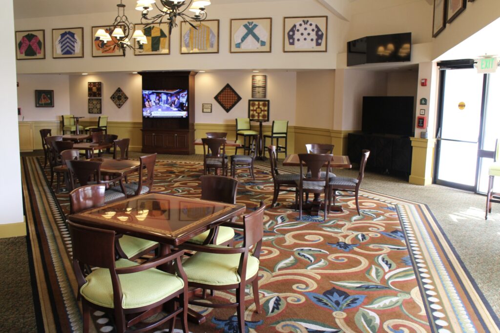 The Turf Club Lounge has a swirly, earth toned carpet with brown tables and chairs, televisions on the walls as well as jockey attire.