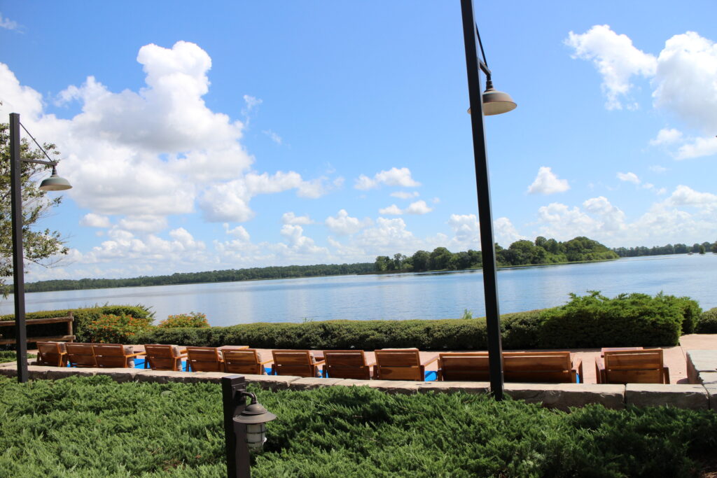 A bright blue sky dotted with fluffy white clouds paints the sky over a reflective blue lake that meets the shore to find a paved seating area and rustic wood chairs behind Wilderness Lodge restaurant Geyser Point