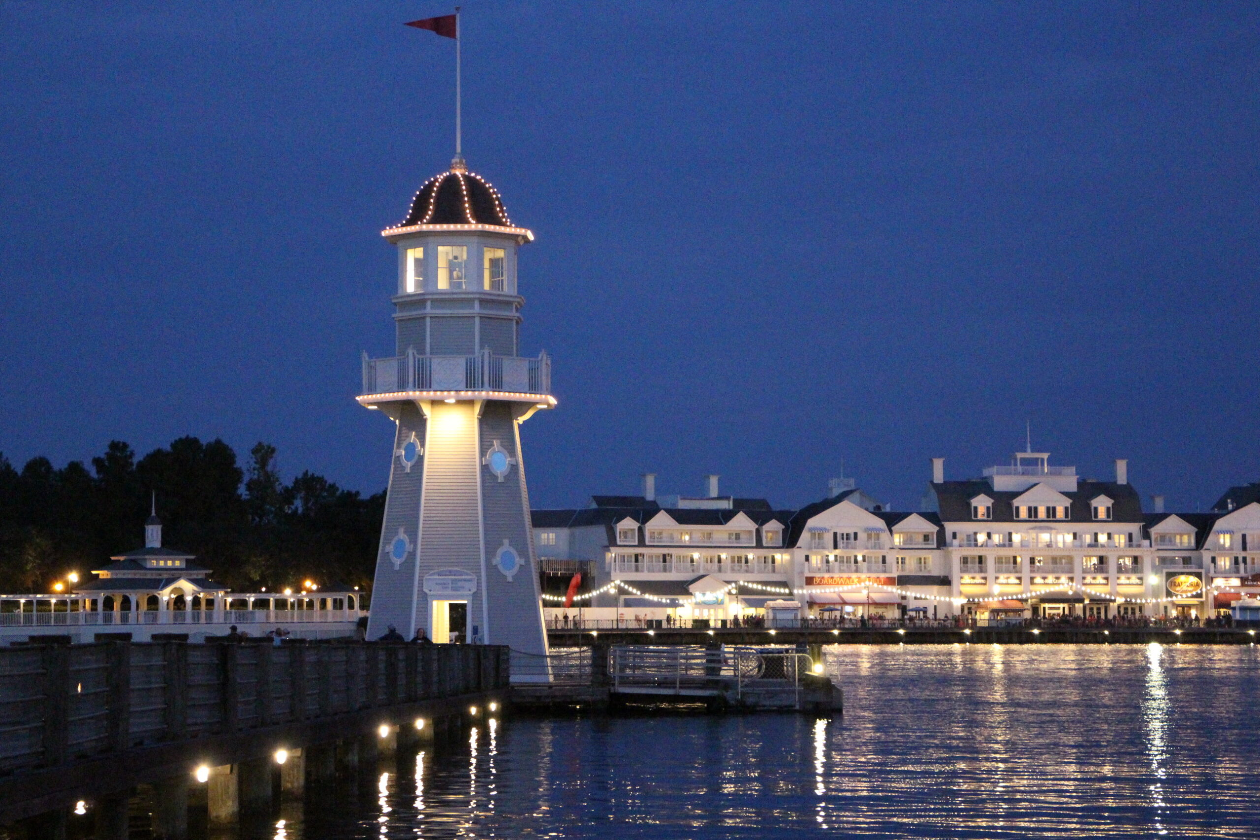 A gray lighthouse sits at the end of a dock with Disney's BoardWalk Resort off in the distance.