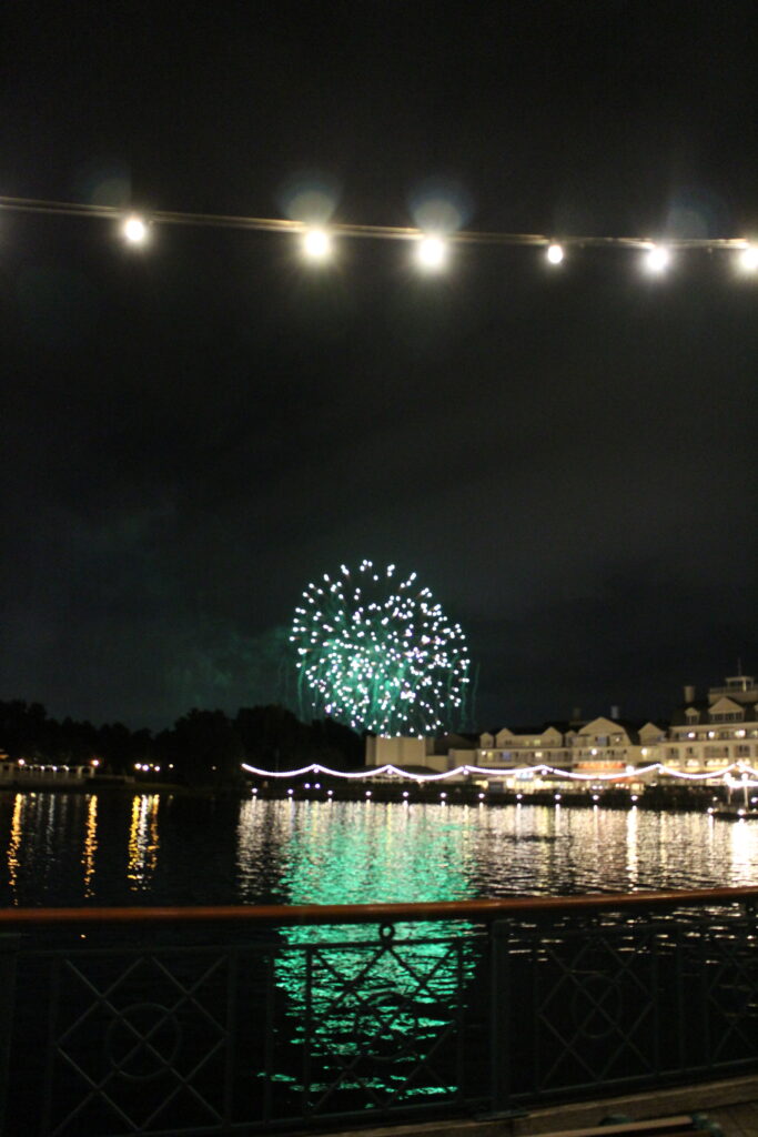 View of fireworks from Disney BoardWalk reflecting over Crescent Lake with string lights and BoardWalk restaurants across the water