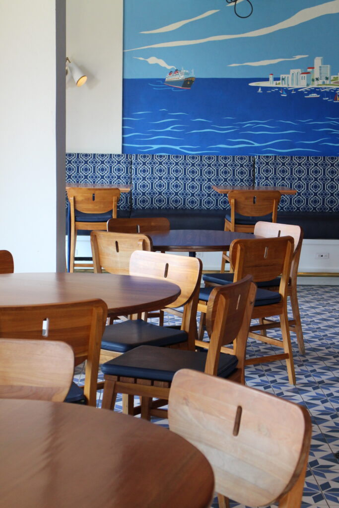 Inside Bar Riva at Rivera Resort wooden chairs sit at round tables of the same color. There are dark shades of blue in the floor tile, wall bench and wall mural.