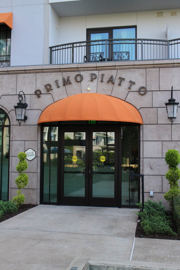 Primo Piatto letters curved above an outside glass door with an orange, rounded covering. This is the exterior entrance to the Riviera Resort restaurant that's a quick service.