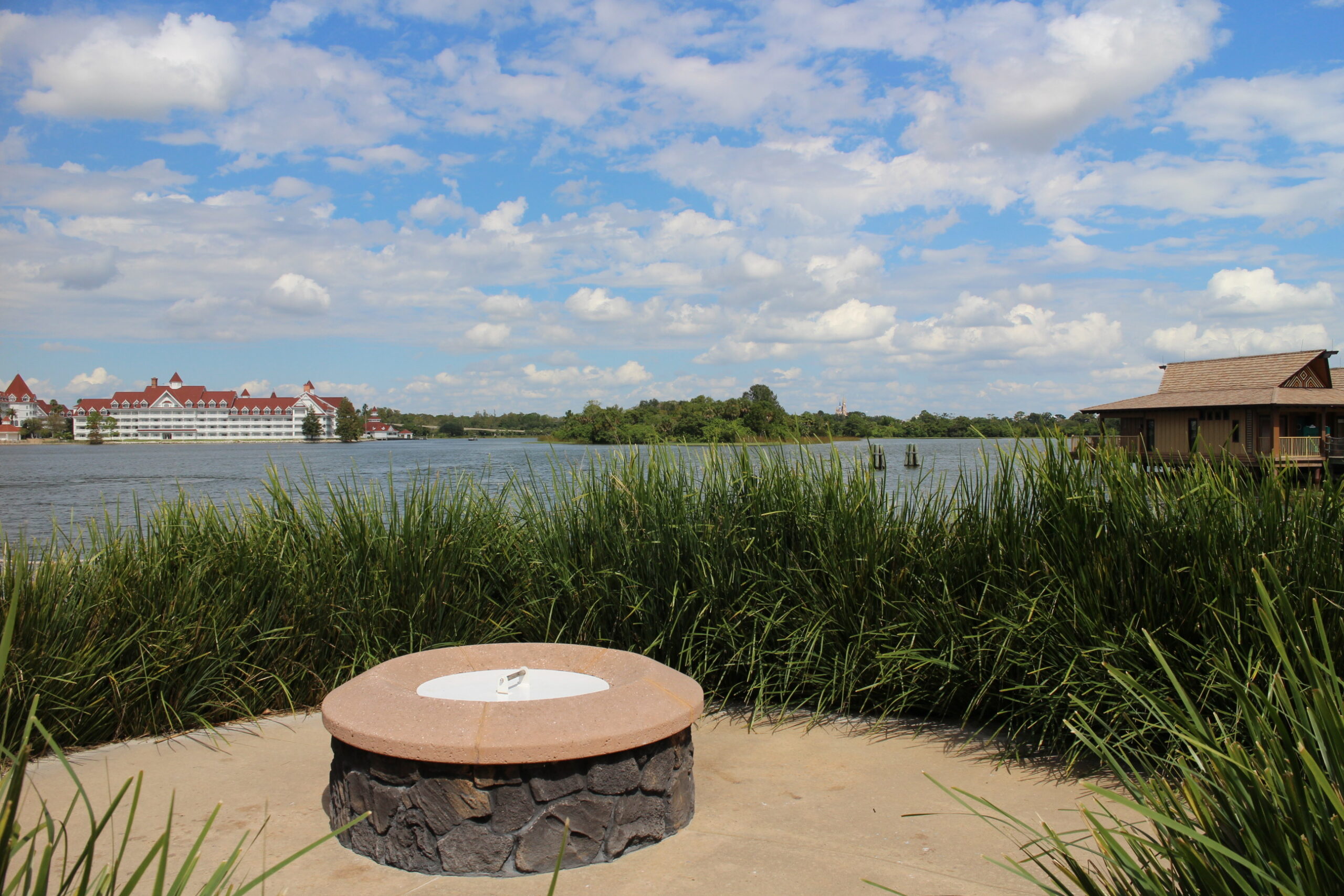 A built in fire pit on the Polynesian Beach surrounded by tall grasses overlooking the lake, blue skies and the Grand Floridian far off in the distance