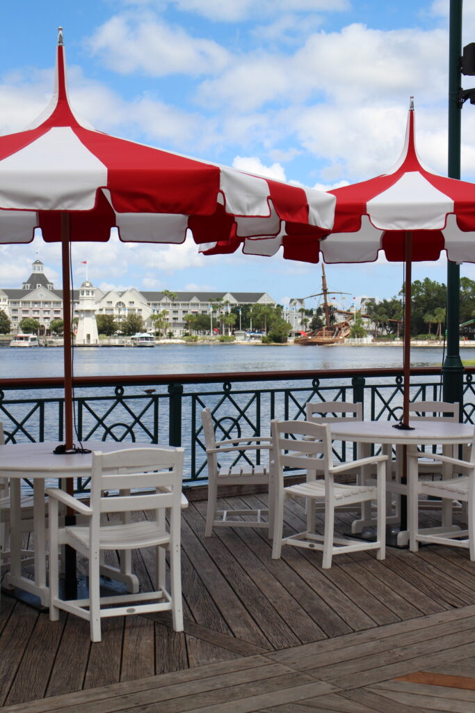 Outdoor seating for Disney BoardWalk restaurants on a wooded deck with white tables and chairs covered by red and white umbrellas