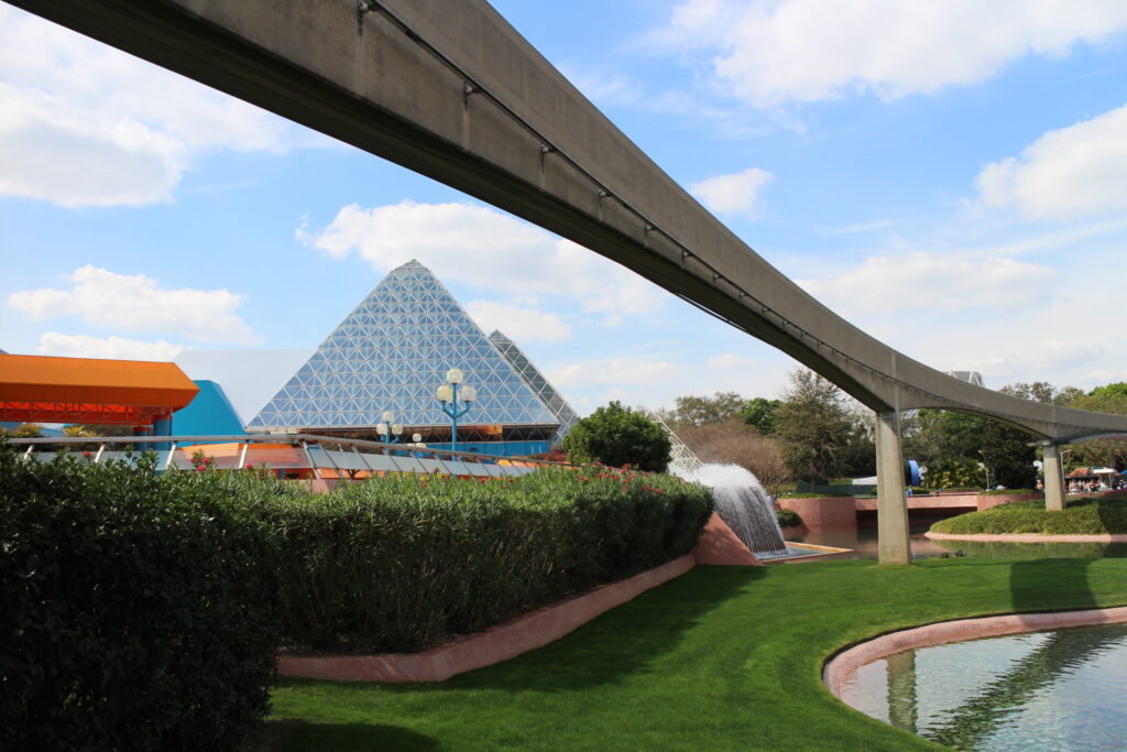 A monorail beam with the Imagination Pavilion off in the distance behind it and green landscaping all around. The Imagination Pavilion is home to Figment, a character that's celebrated at the EPCOT Festival of the Arts.