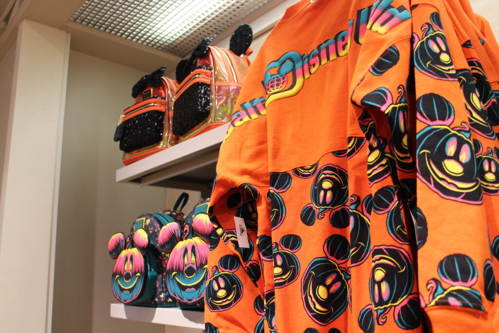 An orange Spirit Jersey with black Mickey pumpkin art hangs next to Halloween Disney Loungefly backpacks
