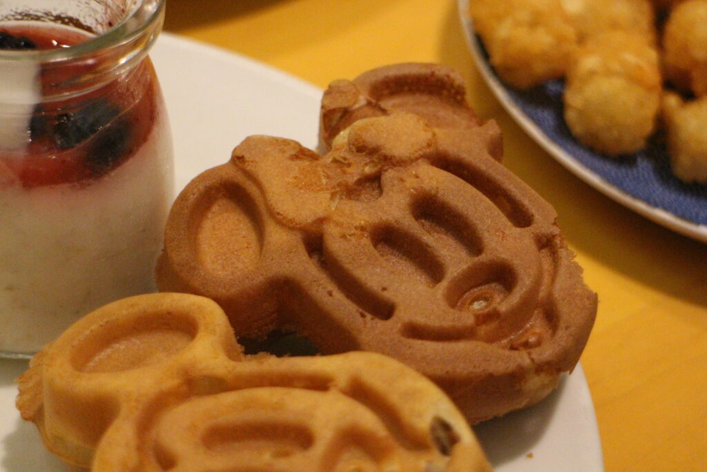 a crisp brown Minnie Mouse miniature waffle on a white plate surrounded by other food that is out of focus