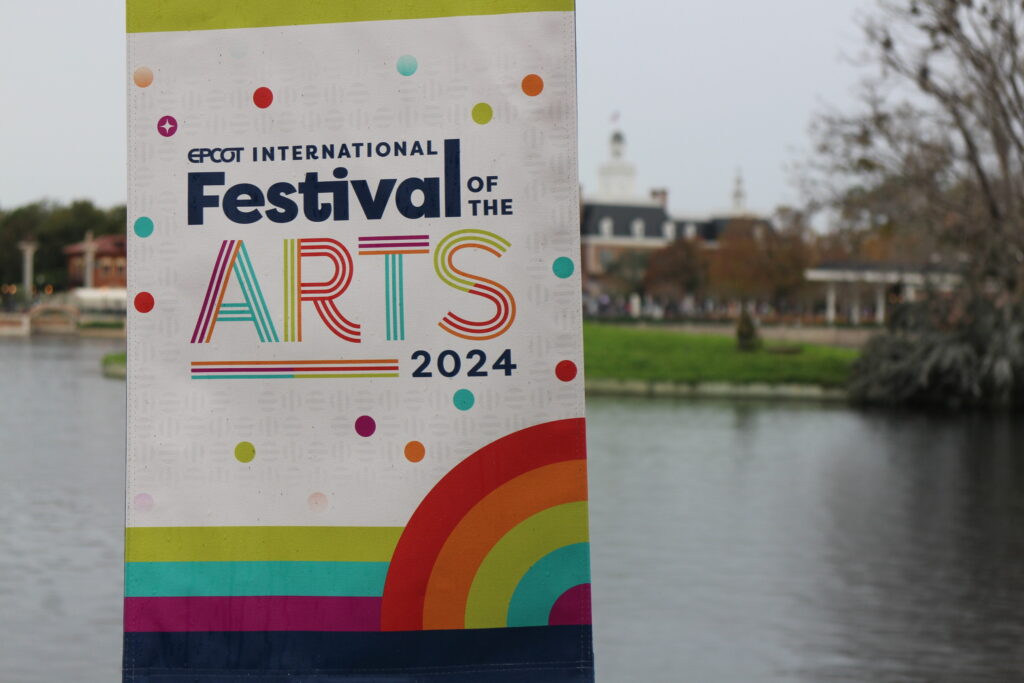 a colorful Epcot arts festival sign stands in front of the large body of water in the theme park