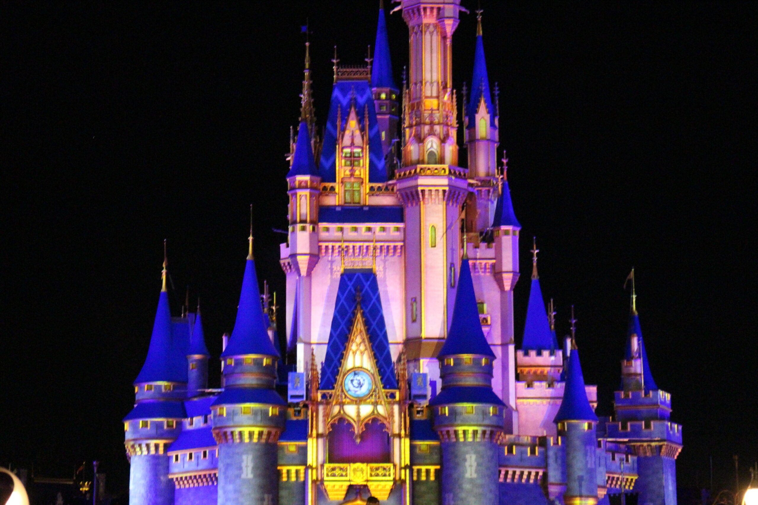 The Disney World castle illuminated at night by projections that make it a vibrant light pink color with turret roofs that are a stunning cobalt blue.