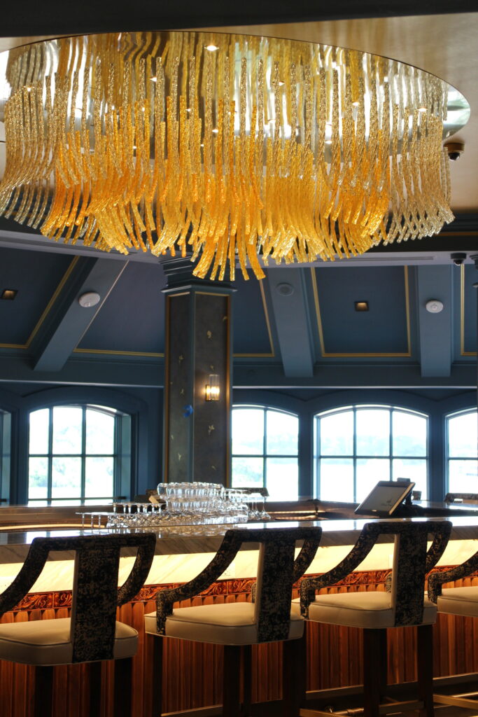 Inside the Enchanted Rose bar and lounge, the bar room has a dark blue-gray walls and ceiling with a bright gold, modern chandelier.