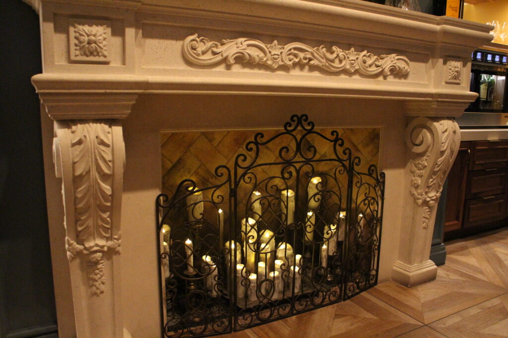 A baroque style white fireplace with candles in place of fire and a black swirly gate in front of them.