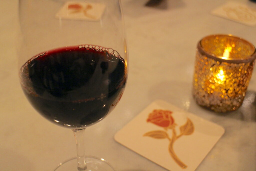 A glass of red wine sits on a table next to a white coaster with a red rose illustration on it and a gold glowing candle holder next to it.
