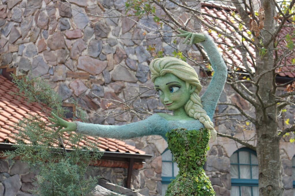 An Elsa topiary does the ice throw pose in front of a stone building.