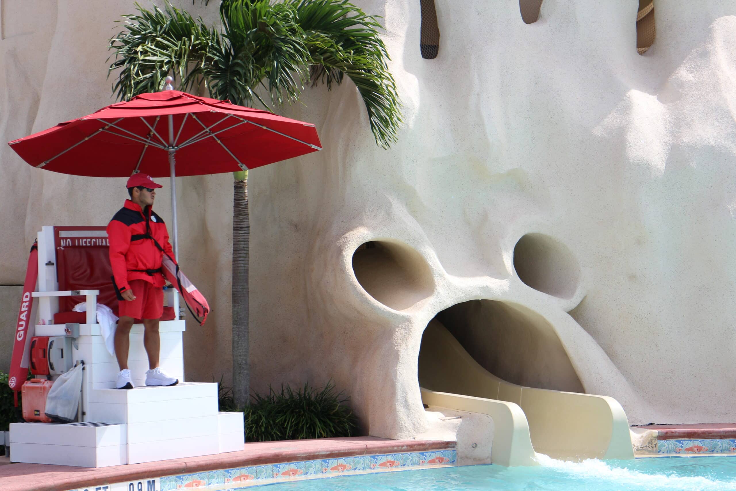 A sandcastle pool at Old Key West that has a Mickey mouse shaped pool slide exit into the pool. A life guard stands to the left in red attire.