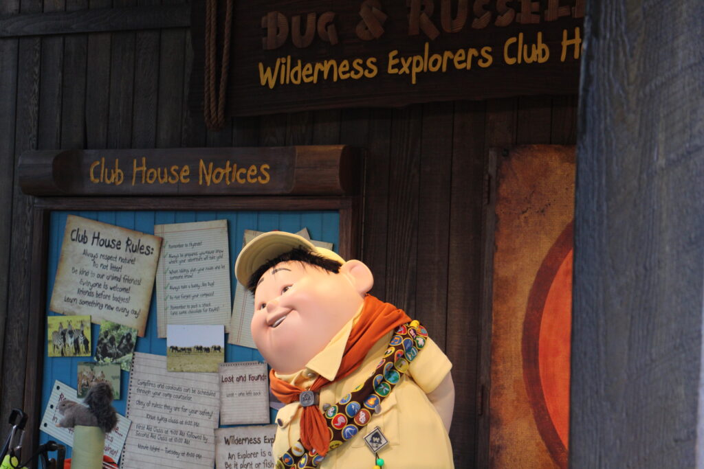 A scout character smiles with his hands behind his back as he waits to greet guests in a covered club house setting. 
