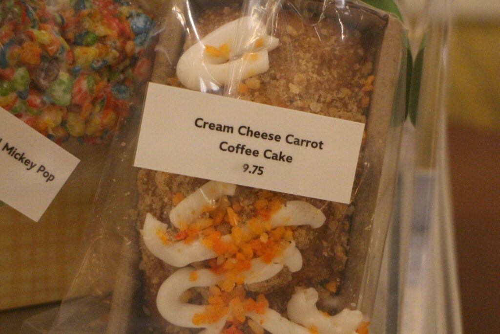 Grand Floridian Cream Cheese Carrot Coffee Cake in a clear wrapper inside a small, rectangular box. The cake has white frosting on top with orange toppings.