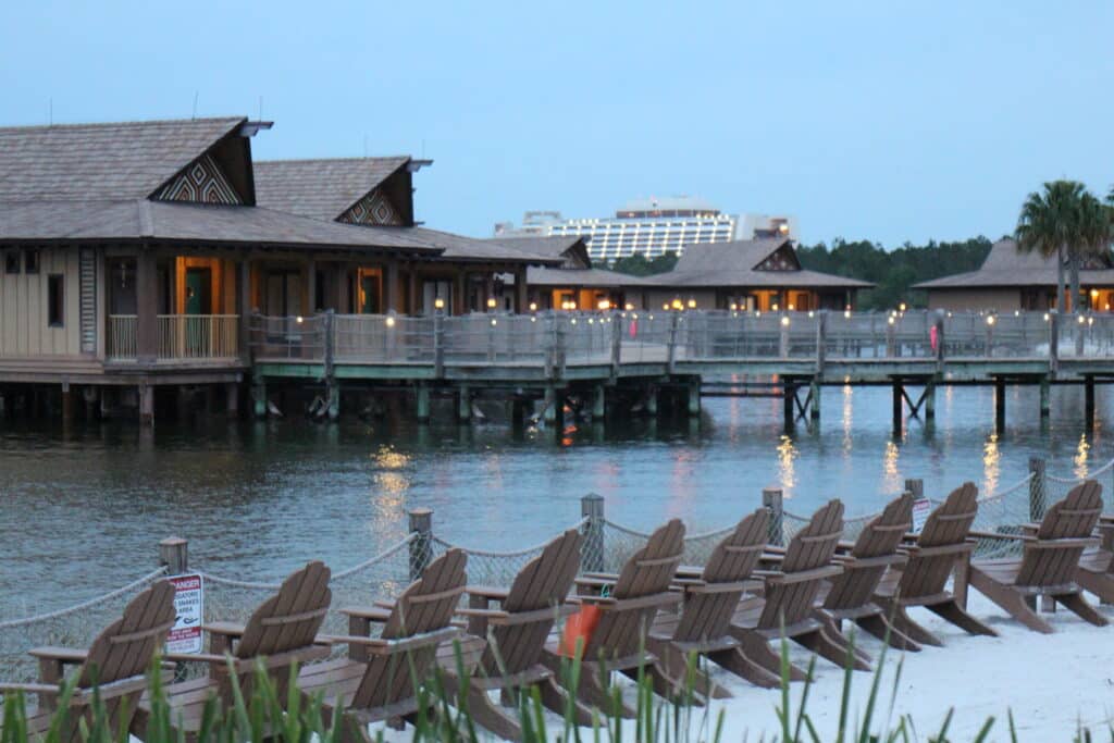 A row of tan Adirondack chairs sits on sugar white sand facing Polynesian bungalows attached to a dock.