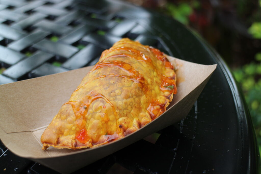 A fried, half moon shaped beef pocket drizzled with an orange sauce in a paper tray on a black table outdoors.