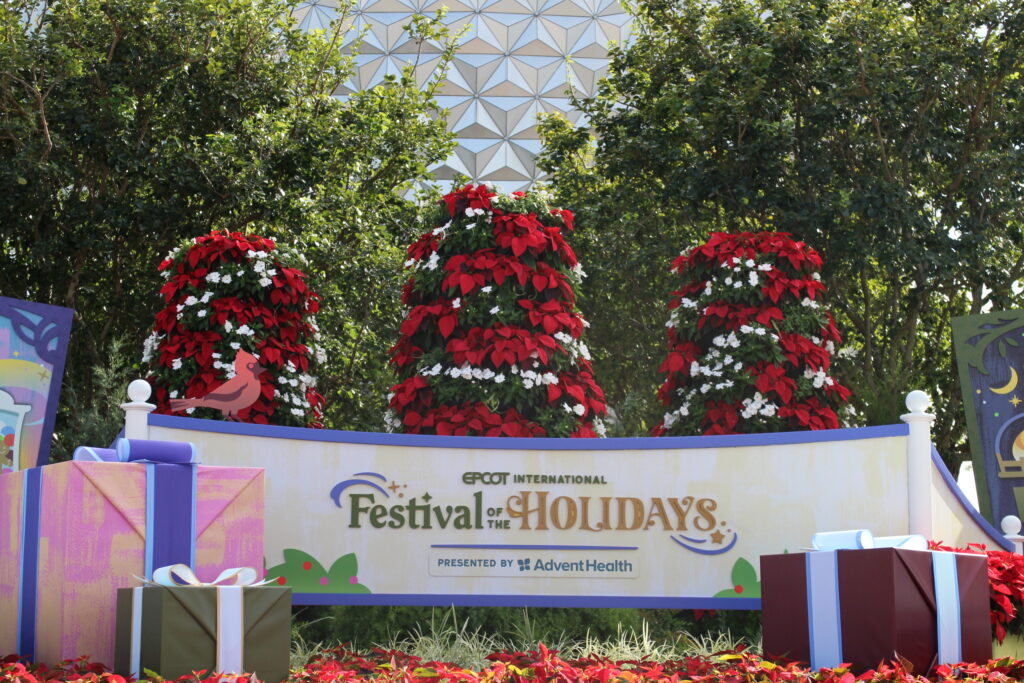 Epcot International Festival of the Holidays Sign at the park entrance with poinsettias and Spaceship Earth behind it.