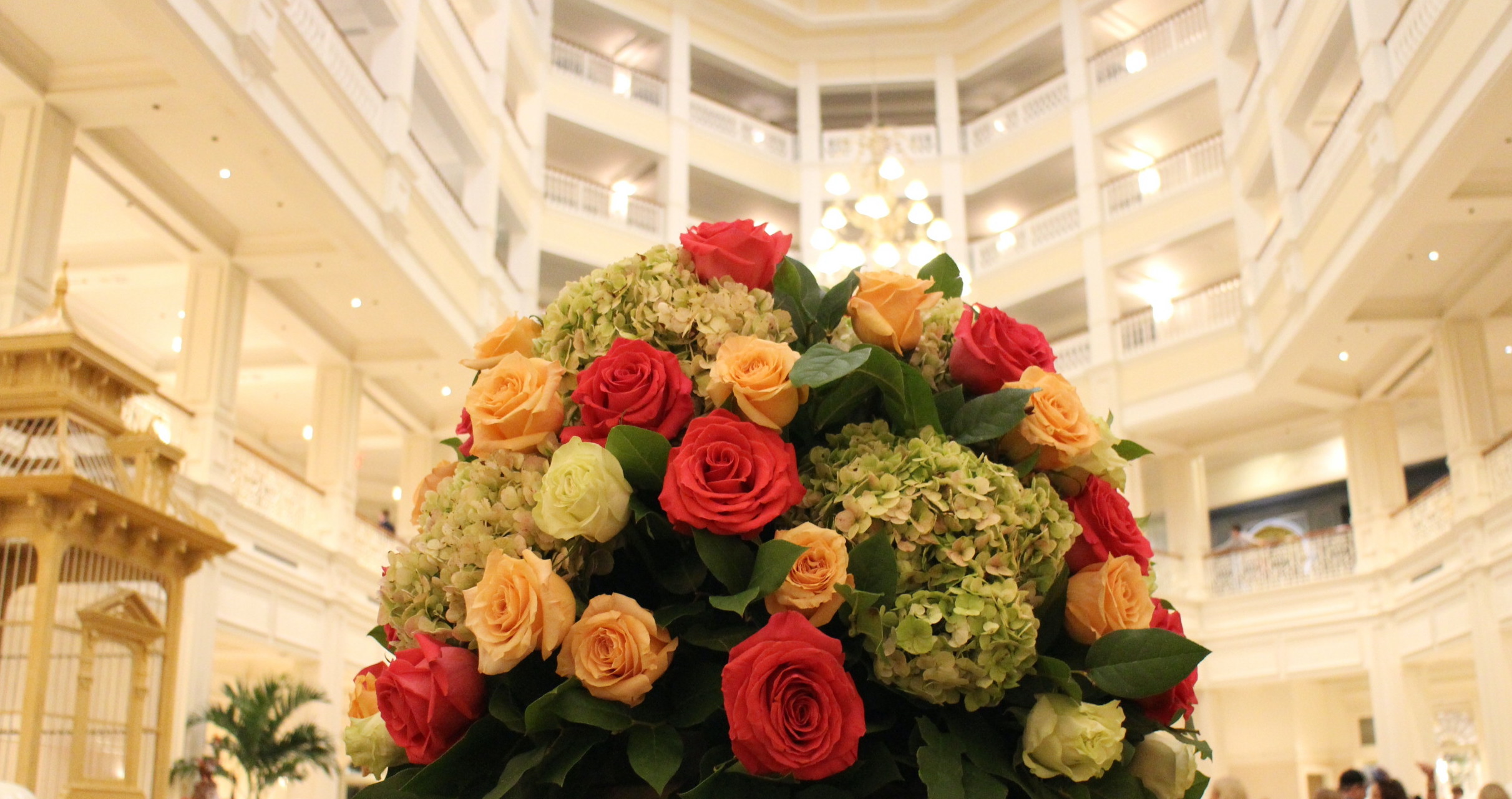 A large floral arrangement featuring roses and hydrangeas with the Grand Floridian resort's bright, multi-story lobby behind it